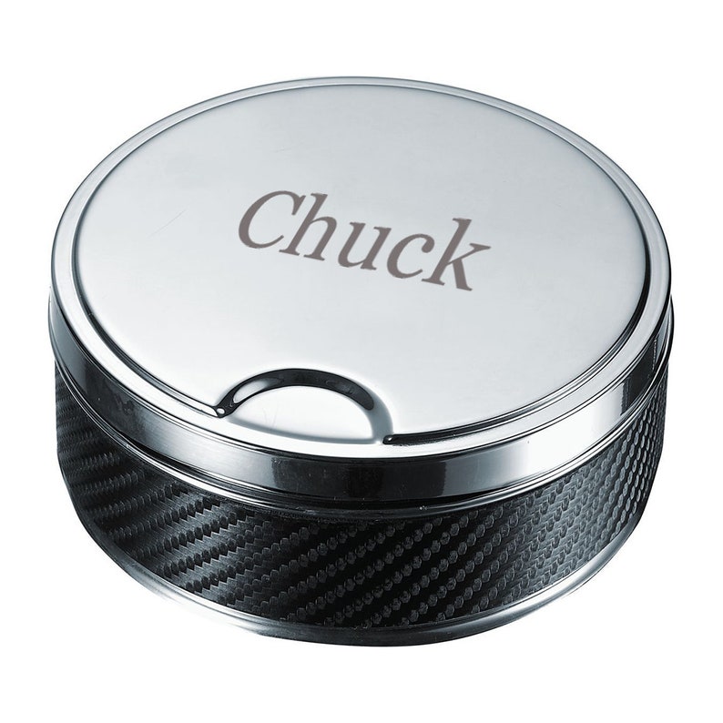 May include: A round, silver-colored ashtray with a carbon fiber pattern on the side. The lid is engraved with the name "Chuck" in a simple font. The ashtray has a small notch on the side for resting items.