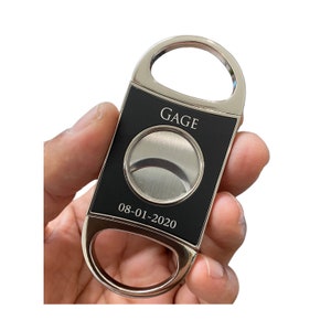 May include: A silver and black cigar cutter with the word "GAGE" and the date "08-01-2020" engraved on it.