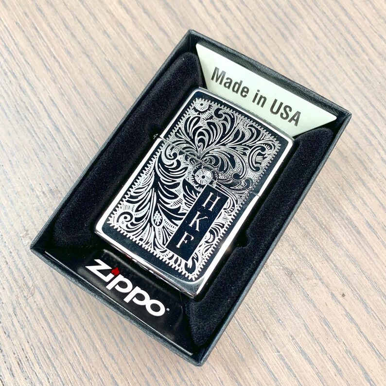 May include: A silver Zippo lighter with a black background and an intricate floral design. The lighter is engraved with the initials "HKF" and the words "Made in USA".