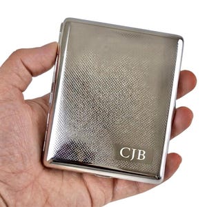 May include: A rectangular silver-toned metal cigarette case featuring a textured crosshatch pattern across the front surface. A smooth, polished rectangular plaque is engraved with the capital letters CJB in a serif font at the bottom right corner.