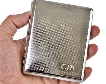 May include: A rectangular silver-toned metal cigarette case featuring a textured crosshatch pattern across the front surface. A smooth, polished rectangular plaque is engraved with the capital letters CJB in a serif font at the bottom right corner.