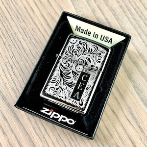 May include: A silver Zippo lighter with a black and white engraved design. The design features a floral pattern with the initials "CEA" in the center. The lighter is in a black box with the words "Made in USA" and "Zippo" printed on it.
