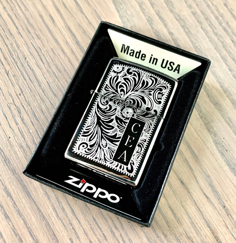 May include: A silver Zippo lighter with a black and white engraved design. The design features a floral pattern with the initials "CEA" in the center. The lighter is in a black box with the words "Made in USA" and "Zippo" printed on it.