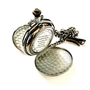 May include: A silver pocket watch with a hinged lid that opens to reveal a space for a photo. The watch has a chain attached to it. The back of the watch has instructions on how to insert a photo: "Turn photo glass until notches are aligned. Tap out glass cover. Insert photo. Align nibs and turn to lock in place."
