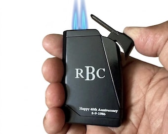 Puede incluir: Un encendedor de metal negro con una llama azul doble que emana de la parte superior. La superficie frontal está grabada con las letras RBC en una fuente serif, seguidas del texto Happy 40th Anniversary y la fecha 8-9-1986 en una fuente blanca más pequeña.