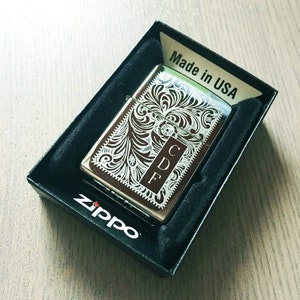 May include: A silver Zippo lighter with an engraved floral design and the initials "CDF" on the front. The lighter is in a black box with the words "Made in USA" and "Zippo" printed on it.