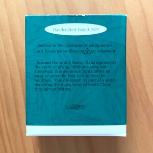 May include: A teal blue box with white trim and a white oval label that reads "Handcrafted-Dated 1995". The box has text that reads "Second in the Centuries of Santa Series. Look for series verification 2 on ornament. Around the world, Santa Claus represents the spirit of giving. With his arms outstretched, this generous Santa offers an array of splendid toys to brighten the holidays. This ornament is part of a series depicting the many faces of Santa Claus throughout history."