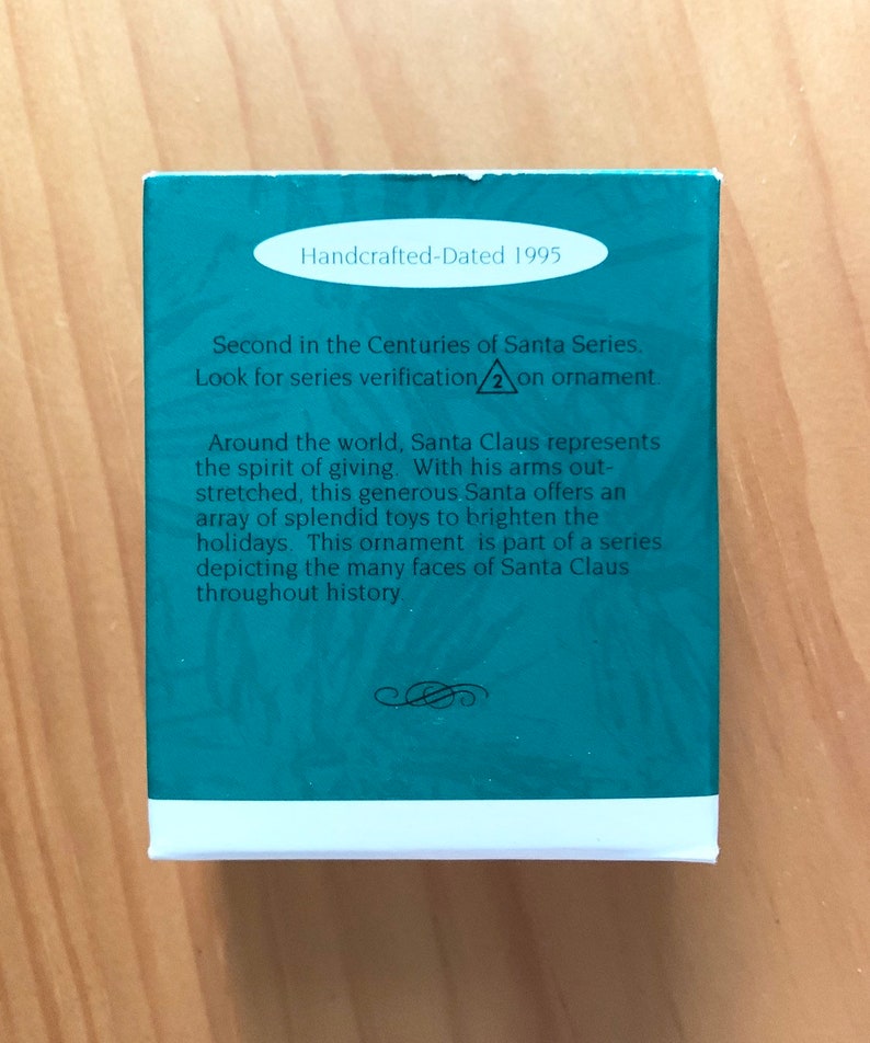 May include: A teal blue box with white trim and a white oval label that reads "Handcrafted-Dated 1995". The box has text that reads "Second in the Centuries of Santa Series. Look for series verification 2 on ornament. Around the world, Santa Claus represents the spirit of giving. With his arms outstretched, this generous Santa offers an array of splendid toys to brighten the holidays. This ornament is part of a series depicting the many faces of Santa Claus throughout history."