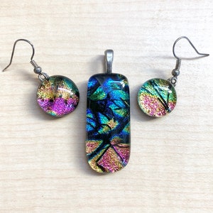 May include: A set of iridescent glass jewelry featuring a pendant and earrings. The pendant is rectangular and the earrings are round. The glass is a mix of blue, green, gold, and pink.