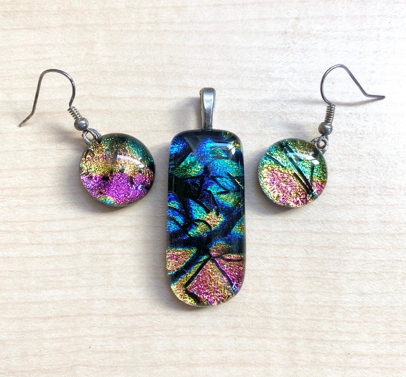 May include: A set of iridescent glass jewelry featuring a pendant and earrings. The pendant is rectangular and the earrings are round. The glass is a mix of blue, green, gold, and pink.