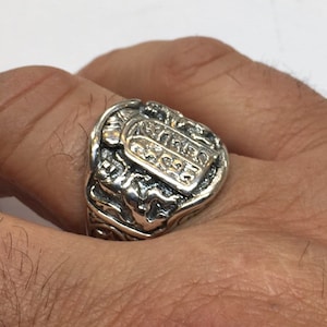 May include: A silver-toned ring featuring a raised rectangular plaque engraved with Hebrew characters. The band displays intricate relief detailing on the sides, including a small figure positioned below the central tablet-shaped emblem.