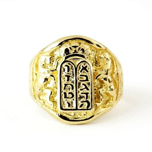 May include: A gold ring featuring a central rectangular plaque engraved with Hebrew characters, topped with a crown motif. The band displays textured, relief-style lion figures on both sides, creating a detailed and ornate appearance in polished yellow gold.