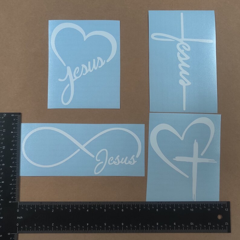 May include: Four white vinyl decals on a light blue background. The decals feature the word "Jesus" in a cursive font with different designs. One decal features the word "Jesus" inside a heart, another features the word "Jesus" inside an infinity symbol, another features the word "Jesus" wrapped around a cross, and the last decal features the word "Jesus" with a cross inside a heart.