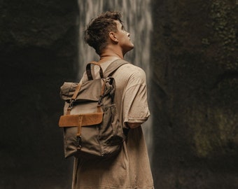 Puede incluir: Un hombre con una camiseta beige y una mochila de lona marrón con detalles de cuero se encuentra frente a una cascada.