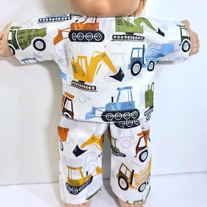 May include: A doll wearing a two-piece pajama set. The pajamas are white with a colorful pattern of construction vehicles, including excavators, bulldozers, and trucks. The doll has blue eyes, a smiling expression, and light brown hair.