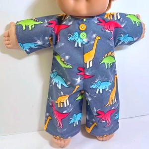 May include: A Cabbage Patch Kid doll wearing a gray dinosaur-print jumpsuit. The jumpsuit features colorful dinosaur illustrations in red, green, blue, and yellow. The doll has blue eyes and light brown hair.