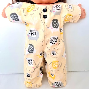May include: A doll wearing a light yellow onesie with a repeating elephant pattern in gray, black, and yellow. The onesie has long sleeves, long pants, and two black buttons. The elephant design is whimsical and playful.