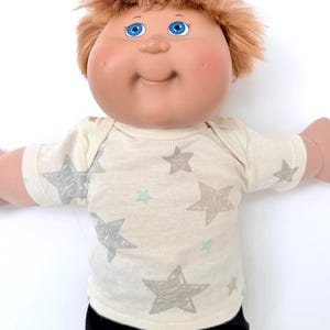 May include: A Cabbage Patch Kid doll with blue eyes and light brown hair. The doll is wearing a cream-colored onesie with gray and light blue star patterns. The doll's arms are outstretched.