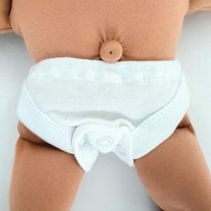 May include: A tan-colored fabric doll torso featuring a prominent belly button and wearing white cotton underwear. The garment is constructed with a wide waistband and a central snap closure detail at the front, providing a snug fit around the doll's midsection.
