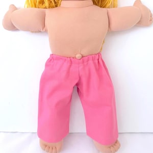 May include: A doll with blonde yarn hair and a light skin tone, wearing bright pink pants. The doll's arms are outstretched, and its feet are visible. The pants have an elastic waistband.