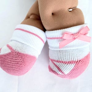 May include: A pair of white and dusty pink baby socks featuring a ribbed cuff, a solid pink toe and heel section, and a small pink satin ribbon bow attached to the side of the cuff on one sock, worn on a pair of soft, tan-colored doll legs.