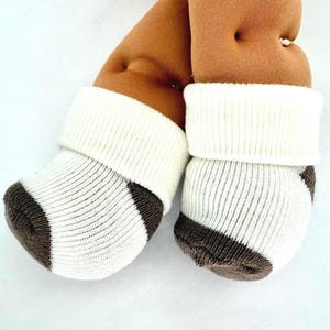 May include: A pair of soft, plush, brown fabric legs with a dimpled texture, wearing white and dark brown striped knit socks with a thick, ribbed white cuff at the top, presented against a plain, light-colored background.