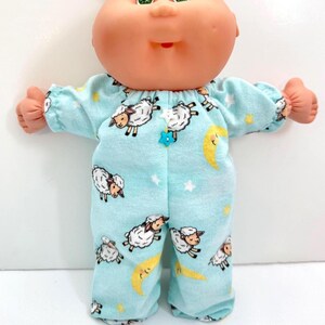 May include: A doll with a round peach-toned head, green eyes, and yarn hair, wearing a light blue long-sleeved romper featuring a repeating pattern of white sheep, yellow crescent moons, and small stars, accented by a small white and blue star charm.