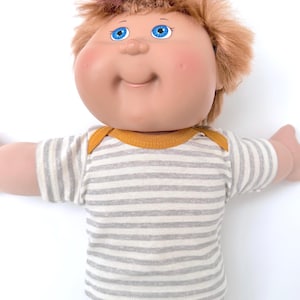 May include: A doll with a round face, bright blue eyes, and messy light brown hair. The doll wears a short-sleeved shirt featuring thin horizontal gray and white stripes with a solid mustard yellow trim around the neckline and shoulder seams.