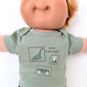 May include: A sage green short-sleeved shirt worn by a doll with orange hair and blue eyes. The shirt features a graphic print with the text MINI EXPLORER, a dinosaur, a volcano, and a small vehicle, all enclosed within dotted square outlines.