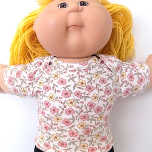 May include: A doll with a round face, brown eyes, and bright yellow yarn hair. The doll wears a short-sleeved shirt featuring a white background with a repeating pattern of small pink and yellow flowers with green leaves, paired with black pants.
