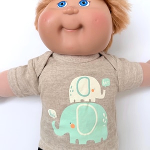 May include: A Cabbage Patch Kid doll wearing a tan onesie with a graphic of two elephants, one light green and one white, stacked on top of each other. The light green elephant has an orange dot and the white elephant has a speech bubble that says "hey".