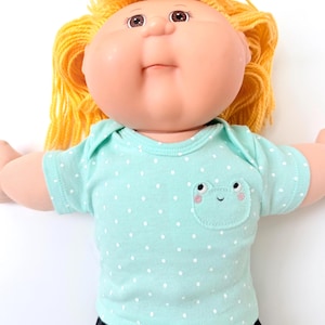 May include: A Cabbage Patch Kid doll with blonde yarn hair and a light skin tone. The doll wears a light teal onesie with white polka dots and a frog pocket. The doll's eyes are brown.