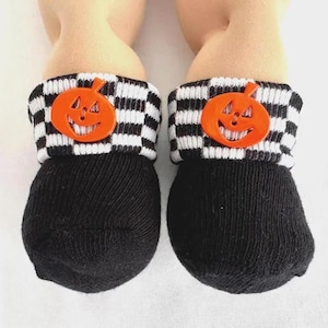 May include: A pair of black socks featuring a white and black checkered cuff. Each cuff is adorned with a bright orange plastic pumpkin-shaped button with a carved jack-o'-lantern face, positioned centrally on the front of the folded fabric.
