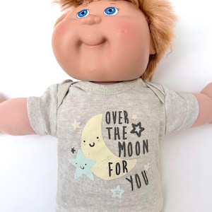 May include: A doll with light skin, blue eyes, and messy brown hair wearing a heather gray short-sleeved shirt. The shirt features a graphic of a smiling yellow crescent moon, a light blue star, and the text OVER THE MOON FOR YOU in black.