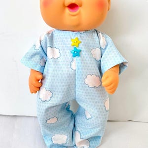 May include: A doll with a round peach-toned head, blue eyes, and rosy cheeks, wearing a light blue one-piece outfit featuring a white cloud pattern and a subtle grid texture. The garment is accented with two small star-shaped buttons in yellow and teal.