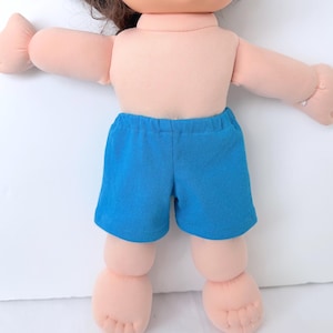May include: A soft-bodied doll with light peach-toned skin and brown hair, wearing bright blue elastic-waist shorts. The doll features a rounded head, outstretched arms, and detailed feet with visible toes, presented in a simple, upright position.