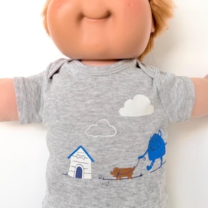 May include: A doll with light brown hair and blue eyes wearing a heather gray short-sleeved shirt. The shirt features a graphic print of a blue monster walking a brown dog near a white doghouse with a blue roof, under two white clouds.