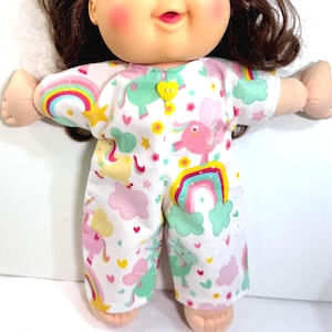 May include: A Cabbage Patch Kid doll wearing a white onesie with a colorful pattern of rainbows, unicorns, clouds, and hearts. The onesie has a yellow heart-shaped button. The doll has brown hair and rosy cheeks.