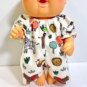 May include: A doll with a round head, rosy cheeks, and blue eyes wearing a cream-colored romper. The romper features a repeating pattern of farm animals, trees, and plants in orange, brown, green, and yellow, with two small buttons at the neckline.