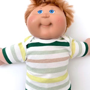 May include: A doll with a round face, rosy cheeks, and bright blue eyes, featuring short, messy reddish-brown hair. The doll is wearing a short-sleeved shirt with horizontal stripes in shades of dark green, light green, yellow, and tan.