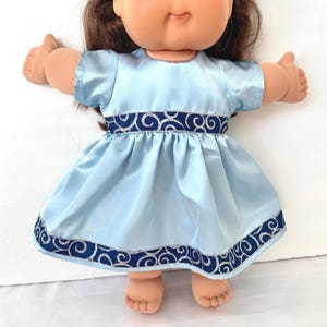 May include: A Cabbage Patch Kid doll wearing a light blue satin dress with short sleeves. The dress features a dark blue band with a silver swirl design at the waist and hem. The doll has brown hair and blue eyes.