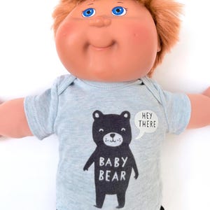 May include: A vintage-style doll with blue eyes and auburn hair, wearing a light blue onesie. The onesie features a black bear graphic with the words "Baby Bear" and a speech bubble that says "Hey There".