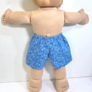 May include: A doll with a light skin tone and soft fabric body wearing blue shorts featuring a repeating pattern of small white, black, and orange triangles. The doll has a rounded belly button, outstretched arms, and visible fingers and toes.