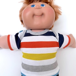 May include: A doll with a round face, rosy cheeks, and bright blue eyes, featuring messy reddish-brown hair. The doll is wearing a short-sleeved shirt with horizontal stripes in navy blue, white, orange, yellow, and light gray, with a gray collar.