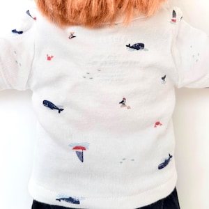 May include: A white short-sleeved t-shirt featuring a repeating pattern of small navy blue whales, red crabs, and sailboats. The garment has a crew neckline and is worn by a figure with reddish-brown hair, showcasing a nautical-themed print design.