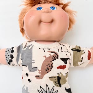 May include: A doll with a round face, bright blue eyes, and messy reddish-brown hair. The doll wears a cream-colored short-sleeved shirt featuring a repeating pattern of various dinosaurs in shades of gray, brown, and olive green, along with small black paw prints.