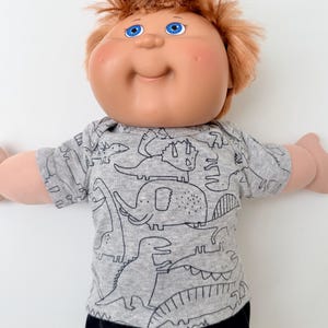 May include: A doll with a round face, rosy cheeks, and bright blue eyes, featuring messy reddish-brown hair. The doll wears a short-sleeved, heather gray crew-neck shirt decorated with a black line-art pattern of various dinosaurs and a mammoth.