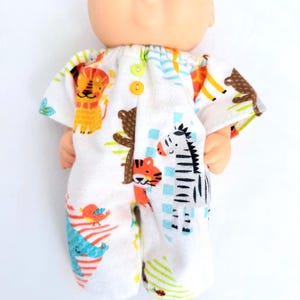 May include: A doll wearing a white short-sleeved romper featuring a colorful print of a lion, zebra, tiger, and other animals. The garment has a gathered neckline and two small yellow buttons at the center front. The doll has a round head and pink cheeks.