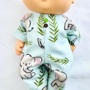 May include: A doll wearing a light blue onesie with a repeating pattern of gray elephants and green fern-like leaves. The onesie has short sleeves and is buttoned up the front with a green and brown button. The doll has rosy cheeks and a smiling expression.