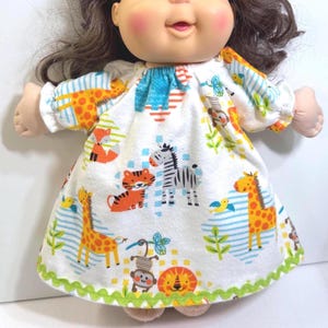 May include: A doll wearing a white dress with a colorful jungle animal print. The dress features giraffes, zebras, tigers, and lions. The dress has a green scalloped trim and blue and orange accents.